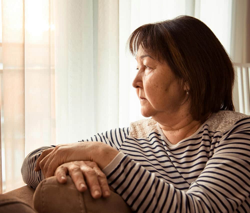 an older woman looks thoughtfully out a curtained window