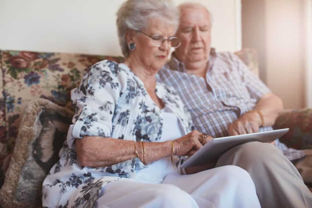 A senior couple sits on their floral sofa- they are looking at something on an ipad together