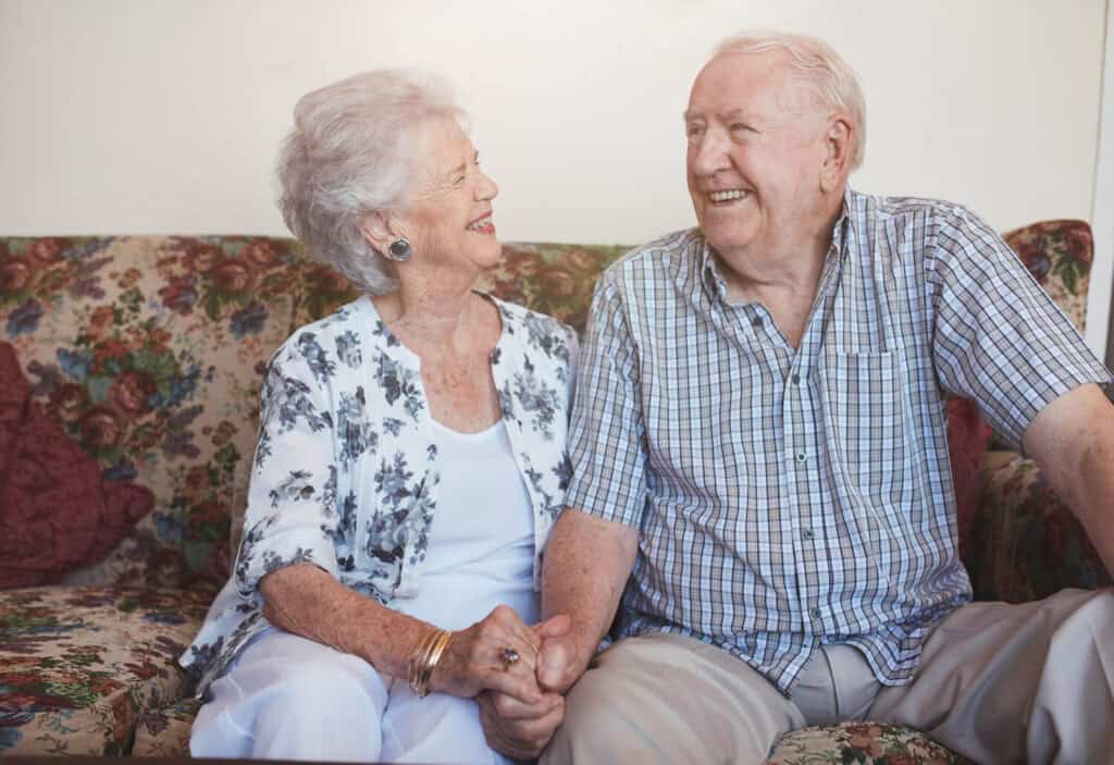 A smiling senior couple seated on their floral sofa look thoughtfully at one another