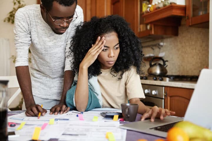 A young woman sits at a kitchen table looking frustrated and tired- important looking paperwork surrounds her and a man looks with concern over her shoulder