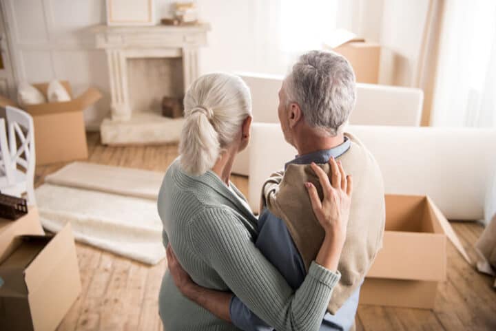 Shot from behind, a man and woman have their arms around each other, looking towards a room in a state of being packed up ready to move