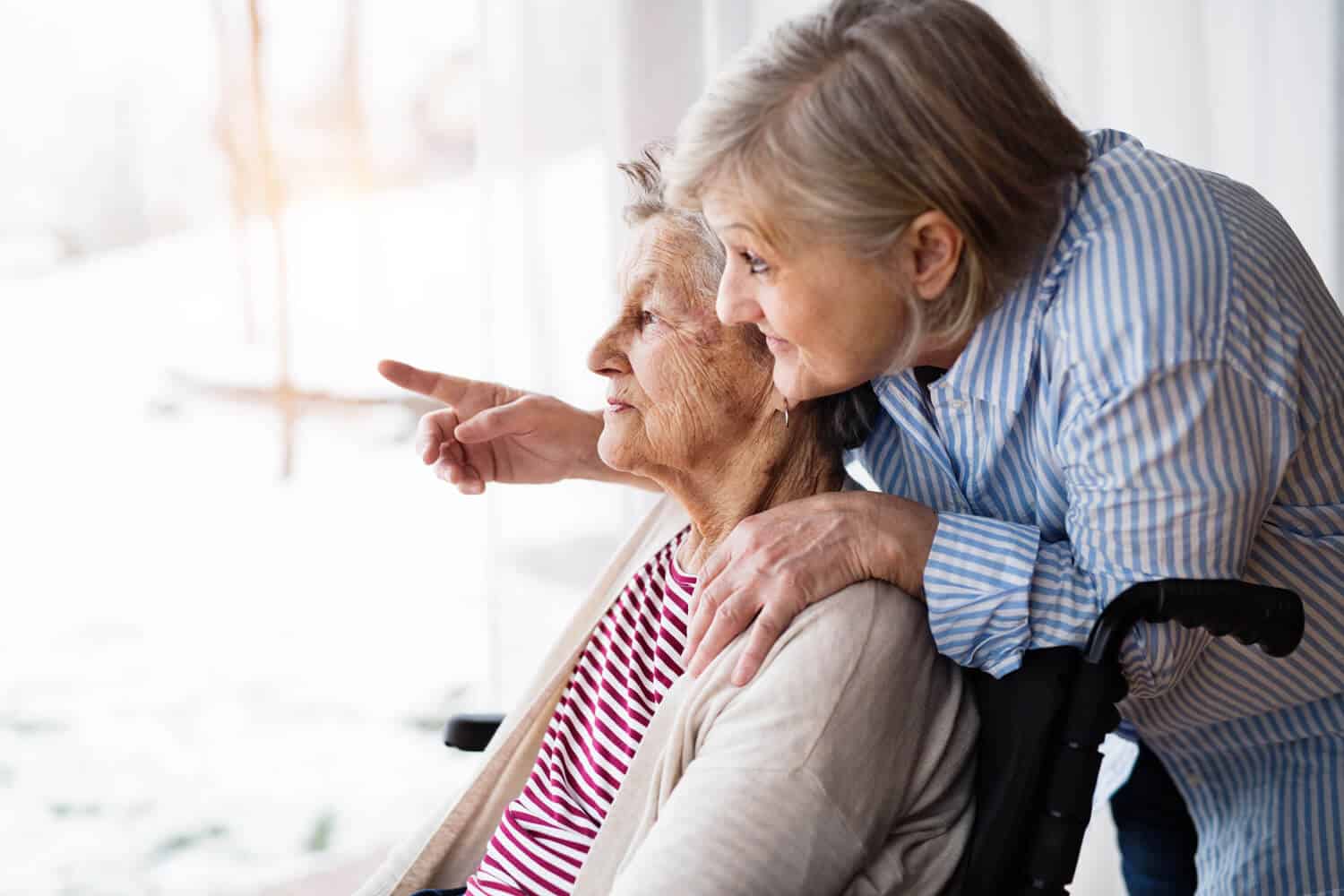 A woman sits in a wheelchair looking out a window- another woman, presumably her daughter, leans over her shoulder lovingly and is pointing at something in the distance
