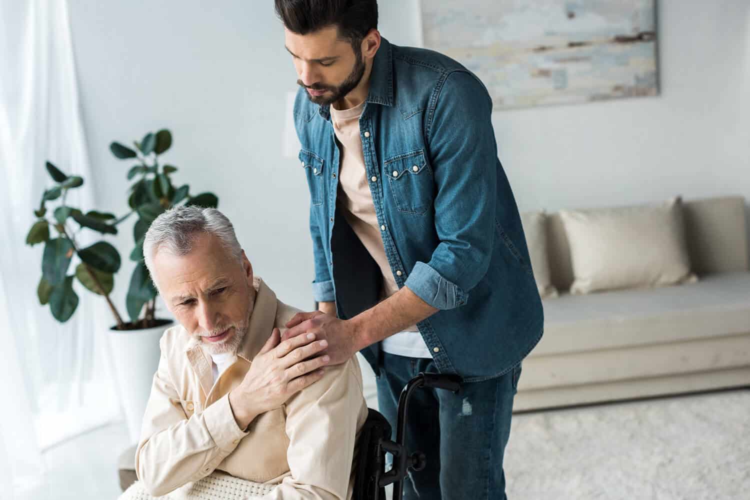 A father and son are together in a simple room- the father sits in a wheelchair, the son has his hand on their shoulder. Both look concerned