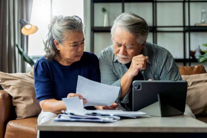 An older couple sits together trying to figure something out- they have confused expressions and they are looking at a pile of paperwork, with their tablet open next to it.