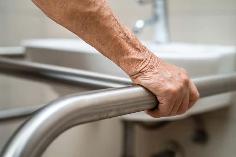 An older person's hand grips a stainless steel support bar in a bathroom setting