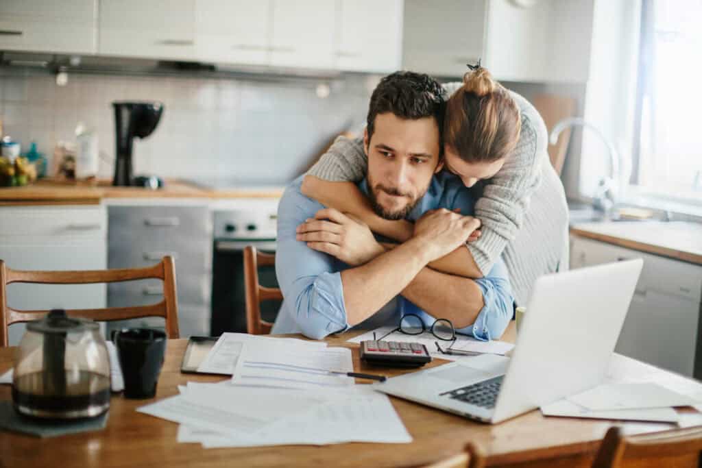 A concerned young man is sitting at a kitchen table strewn with paperwork- a woman lovingly embraces him from behind