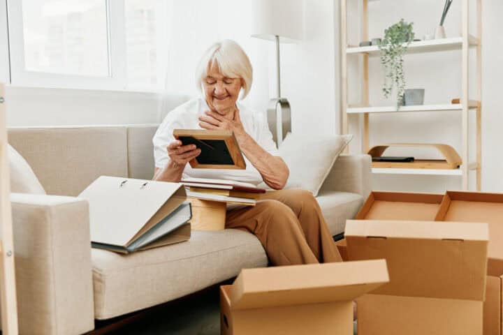 An older woman is sitting on a sofa surrounded by moving boxes in varying states of unpack. She has a pile of photos and is holding one with a warm smile, as if remembering a long ago memory