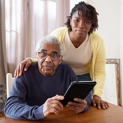 An older man and a wonder woman, presumably his granddaughter, look into the camera. He is seated at a table holding an ipad, and she is standing behind him with a caring hand on his shoulder.