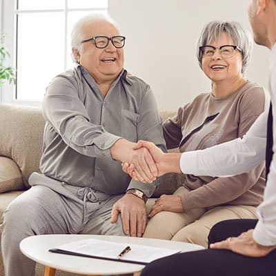 An older man and woman are sitting on a couch across from a professional looking person. The older man is shaking hands warmly with the professional, everyone is smiling