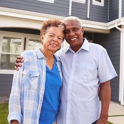 An older smiling couple stands outside in front of a blue house