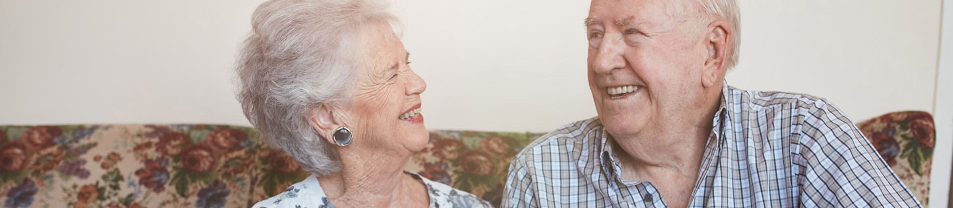 A smiling senior couple seated on their floral sofa look thoughtfully at one another