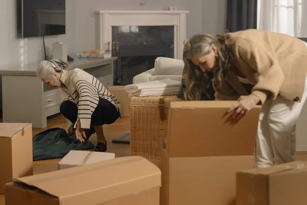 2 women are bent over moving boxes in a living room, focused on their task of packing