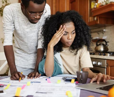 A young woman sits at a kitchen table looking frustrated and tired- important looking paperwork surrounds her and a man looks with concern over her shoulder