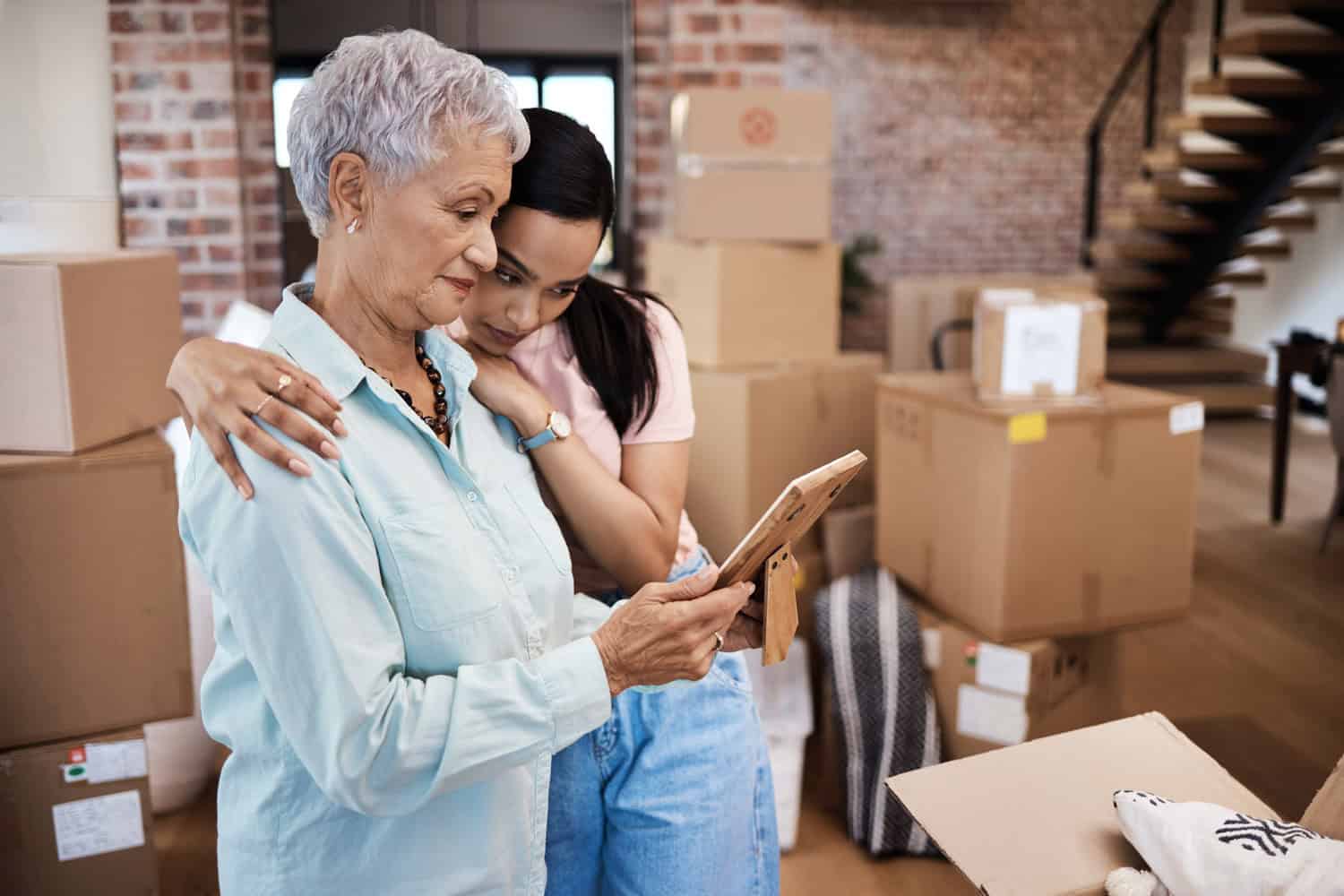 A mother and daughter embrace warmly. The mother holds a photo frame, and both are looking thoughtfully at the image. Moving boxes can be seen stacked up behind them