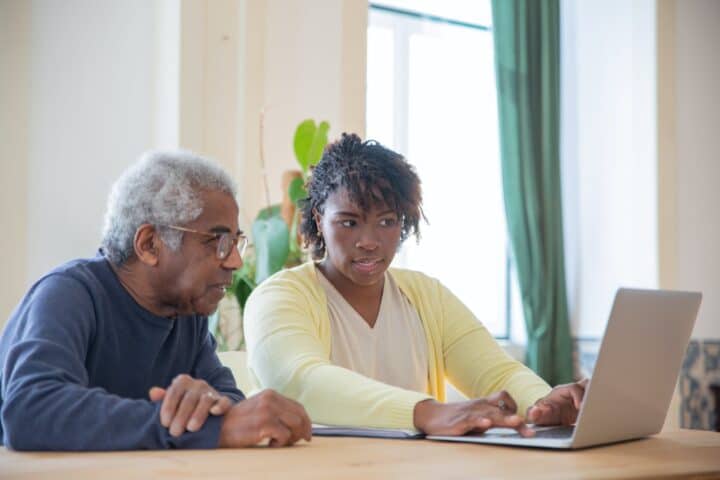 A woman sits at a table with an older man who appears to be a relative- they are looking at something on a laptop together